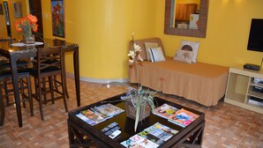 Quarto com cama, mesa de jantar com cadeiras, mesinha de centro com revistas e uma pequena planta.