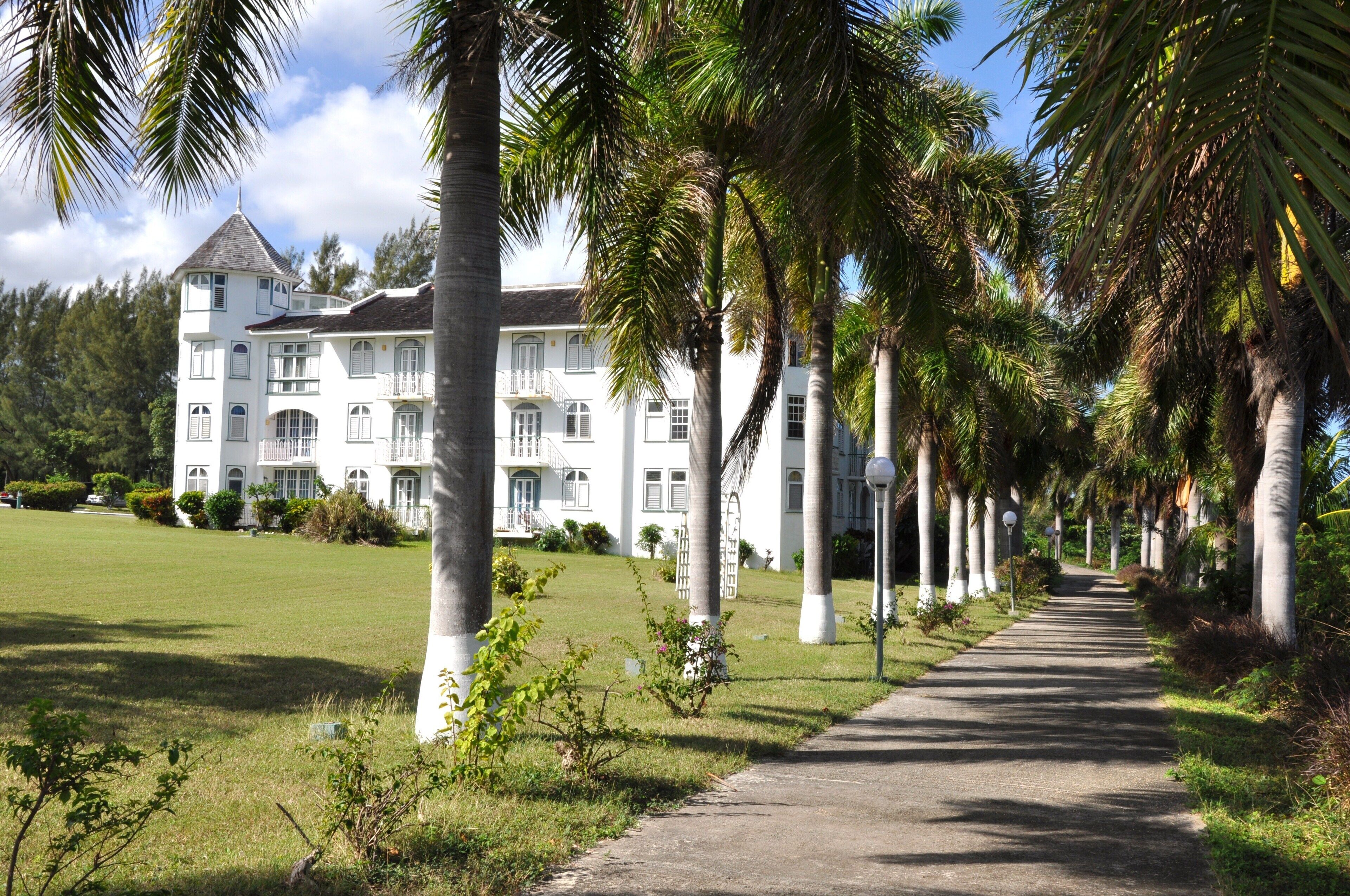 Un grand bâtiment de style colonial, de couleur blanche, doté de nombreux balcons et d’une tour centrale, entouré d’une pelouse bien entretenue et de palmiers.