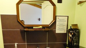 A bathroom with a beige sink, a mirror above it, a trash can, and a shelf with toiletries.