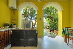 A bright yellow room with a wicker chair, a dining table with a green and white striped tablecloth, and a view of a garden through an archway. - Villa Veronica Villasimius code IUN P5506 (Villasimius)