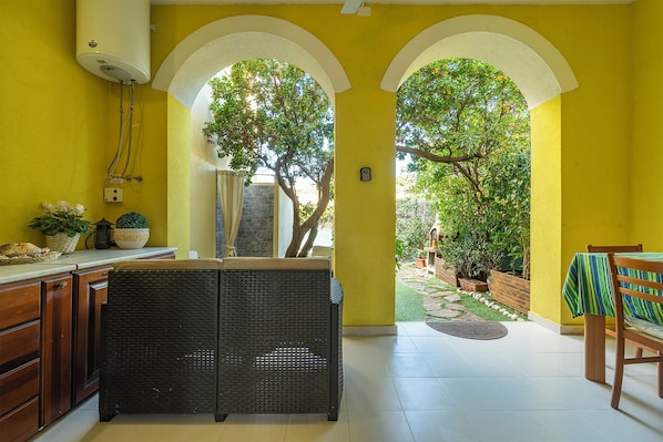 A bright yellow room with a wicker chair, a dining table with a green and white striped tablecloth, and a view of a garden through an archway.