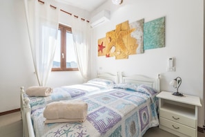 A bedroom with two beds, a window with curtains, wall art, a bedside table with a lamp, and a thermostat.
