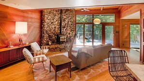 Un salon chaleureux avec une cheminée en pierre, des murs en bois et de grandes fenêtres donnant sur l’extérieur.