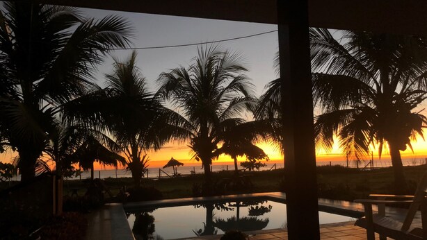 Vista al atardecer desde un espacio al aire libre techado con piscina, palmeras y un patio con baldosas.