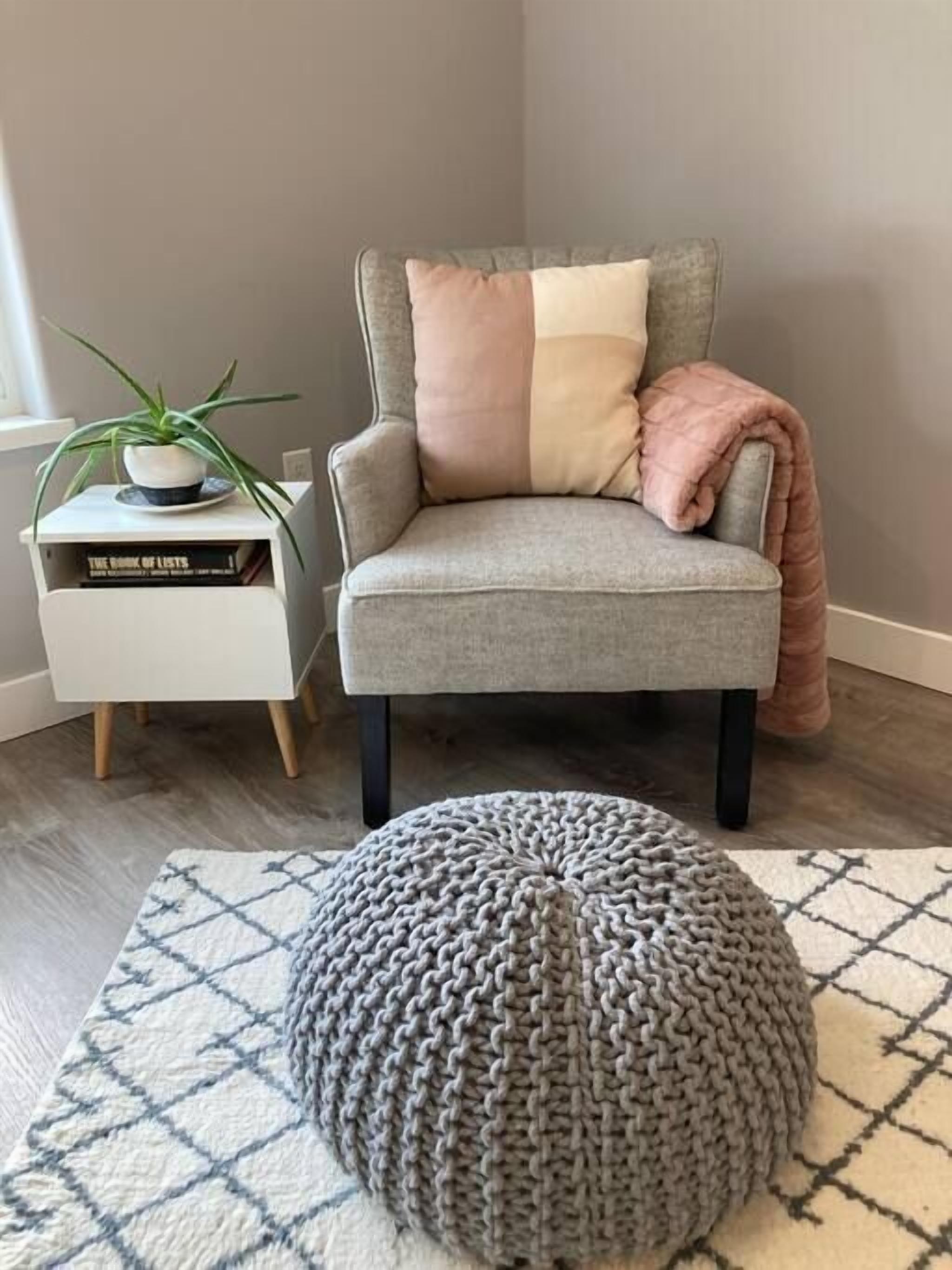 Uma poltrona cinzenta com almofada bege e rosa, uma mesa lateral branca com uma planta e livros, e um banquinho de malha cinzento sobre um tapete estampado.