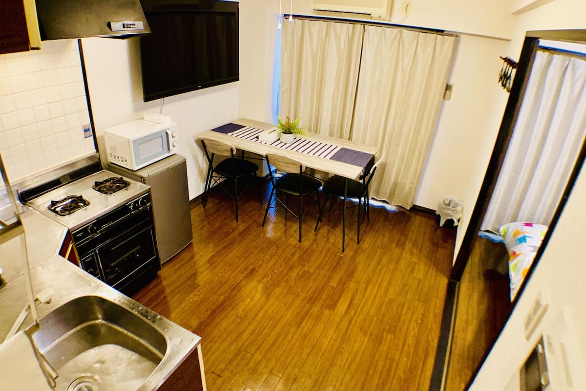 Cocina compacta con estufa, microondas y lavamanos. Hay una mesa de comedor con sillas y una pequeña planta en maceta.