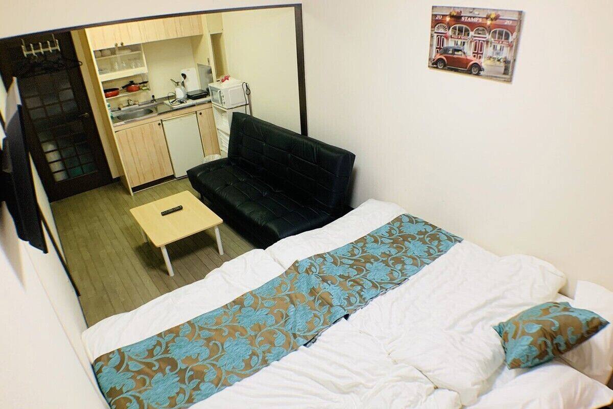 Una habitación pequeña y funcional con una cama, un sofá negro, una mesita y una kitchenette al fondo.