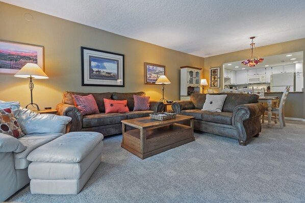 A living room with a sofa, ottoman, and coffee table, a kitchen area with cabinets and a chandelier, and framed pictures on the walls. - Cozy Prime Main St Corner/Steps to Town Lift/Hot Tub Pool/Free Parking/W/D (Park City)