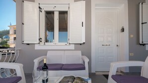 Un balcón con mobiliario de mimbre, una puerta y una ventana con contraventanas.