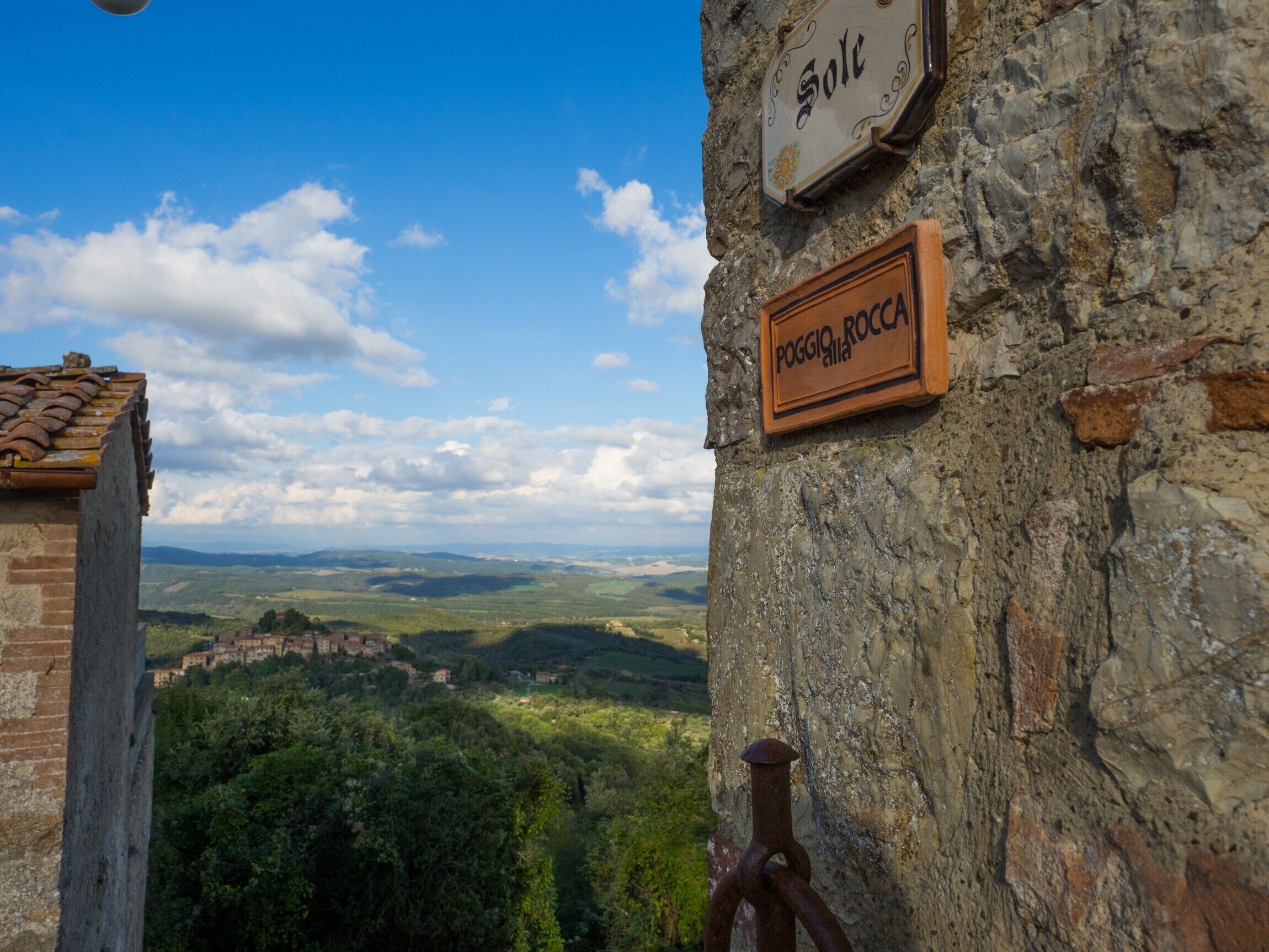 A rustic stone wall with a wooden sign reading "POGGIO ROCCA" and a metal plaque with the word "Sole".