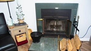 Ein Zimmer mit Kamin, einer schwarzen Ledersofa, einem kleinen Weihnachtsbaum und einem Korb auf einem Holzkabinett.