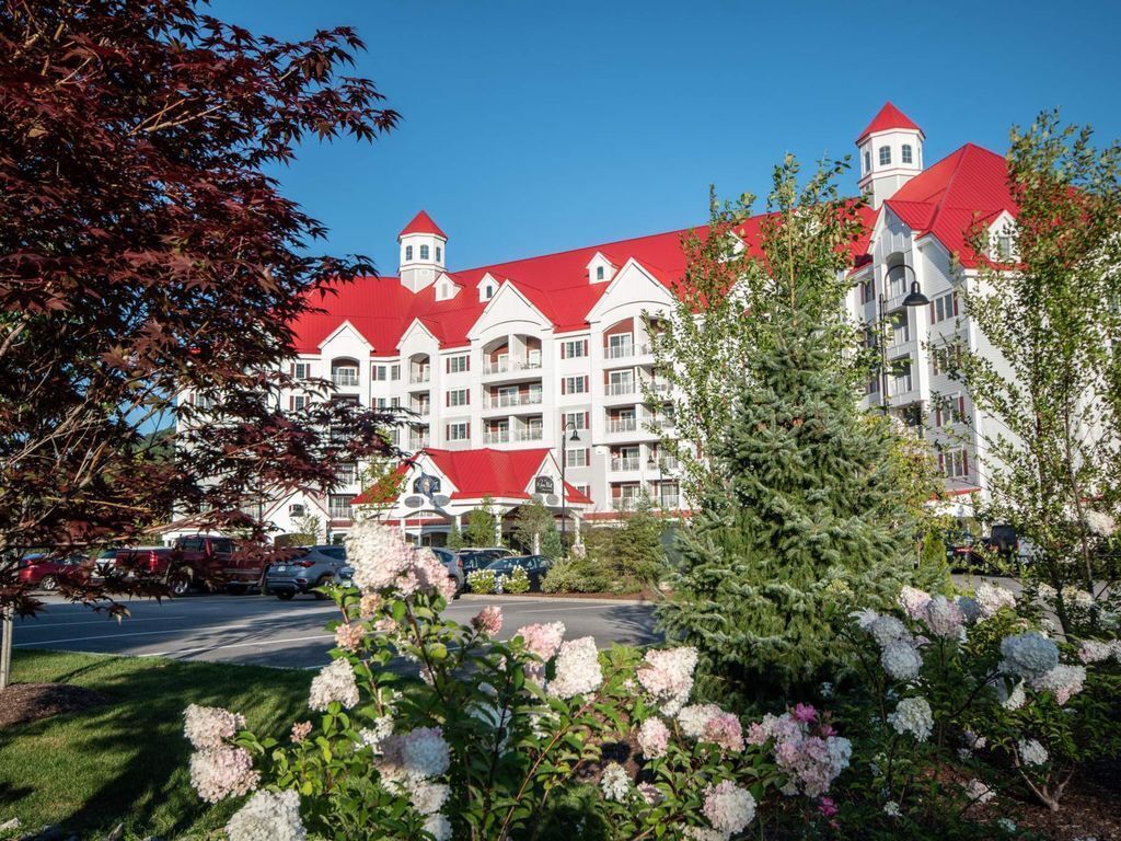 A large hotel with multiple red roofs and white walls, surrounded by lush greenery and flowering plants.