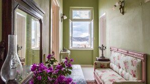 Habitació amb un mirall gran, un vas amb flors morades, un sofà amb tapisseria floral i vista al mar per una finestra.