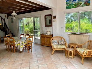 A dining area with a table and chairs, a wicker sofa, and a view of greenery through the windows.