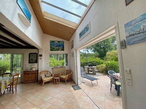 A sunlit room with a dining area, a seating area with chairs, a table, and a view of greenery outside. - Cottage with an idyllic garden at the lake G0239 (Lacanau)