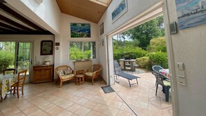 A spacious patio with a dining area, a seating area with a table and chairs, and a view of the garden through large windows. - Renovated landaise style house with enchanted garden, 2 steps from the lake (Lacanau)