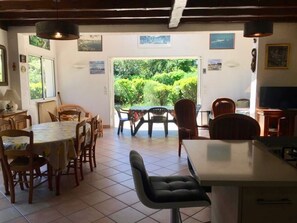 A dining area with a table and chairs, a kitchen area with a counter and appliances, and a view of greenery through an open door. - Cottage with an idyllic garden at the lake G0239 (Lacanau)