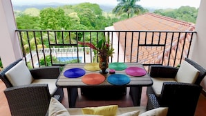 Ein Balkon mit Korbmöbeln, einem Tisch mit bunten Unterlagen und Blick auf ein Schwimmbad und viel Grün.