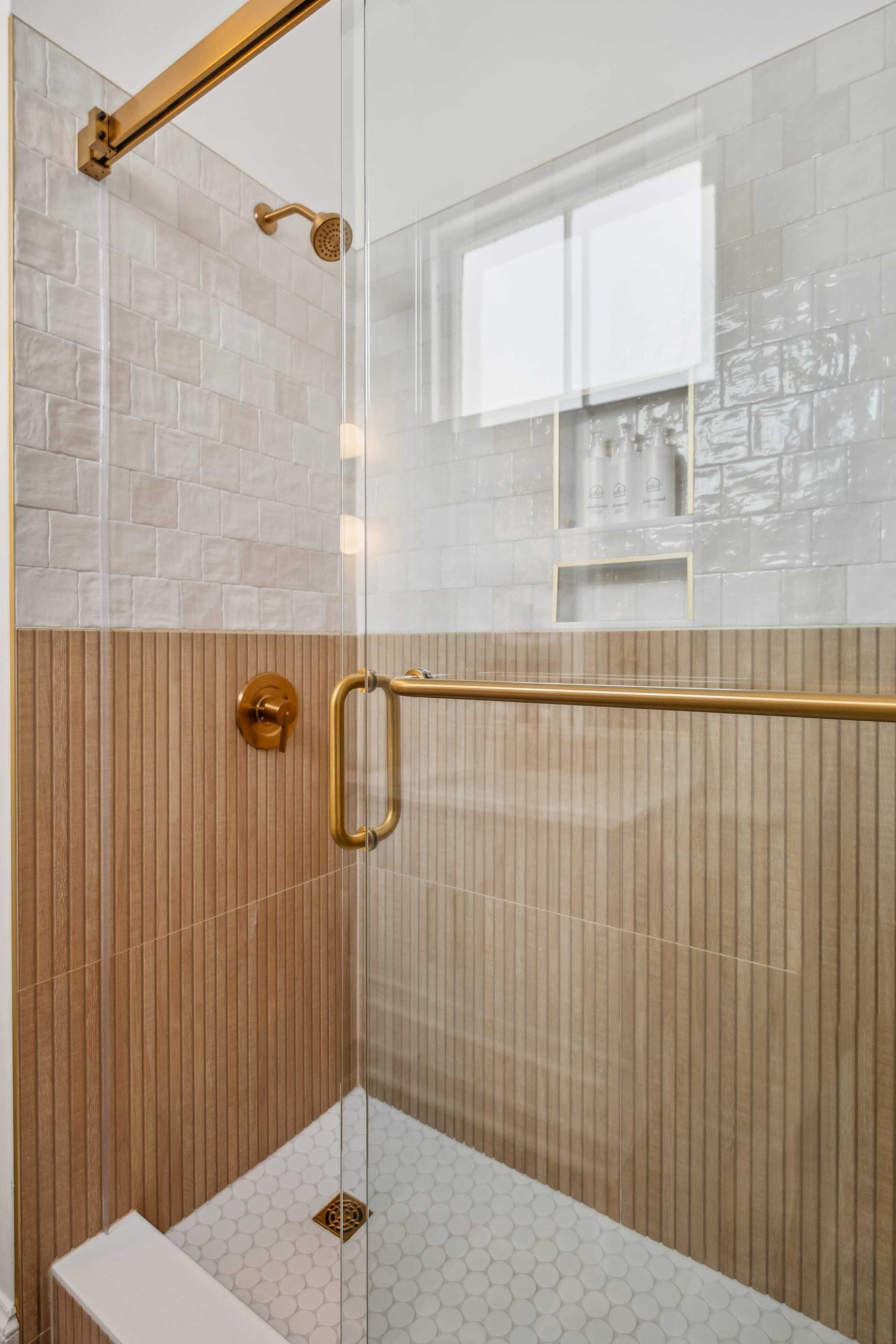 Ducha con cerramiento de vidrio, pared de paneles de madera, ducha con cabezal dorado y piso cubierto de azulejos blancos.