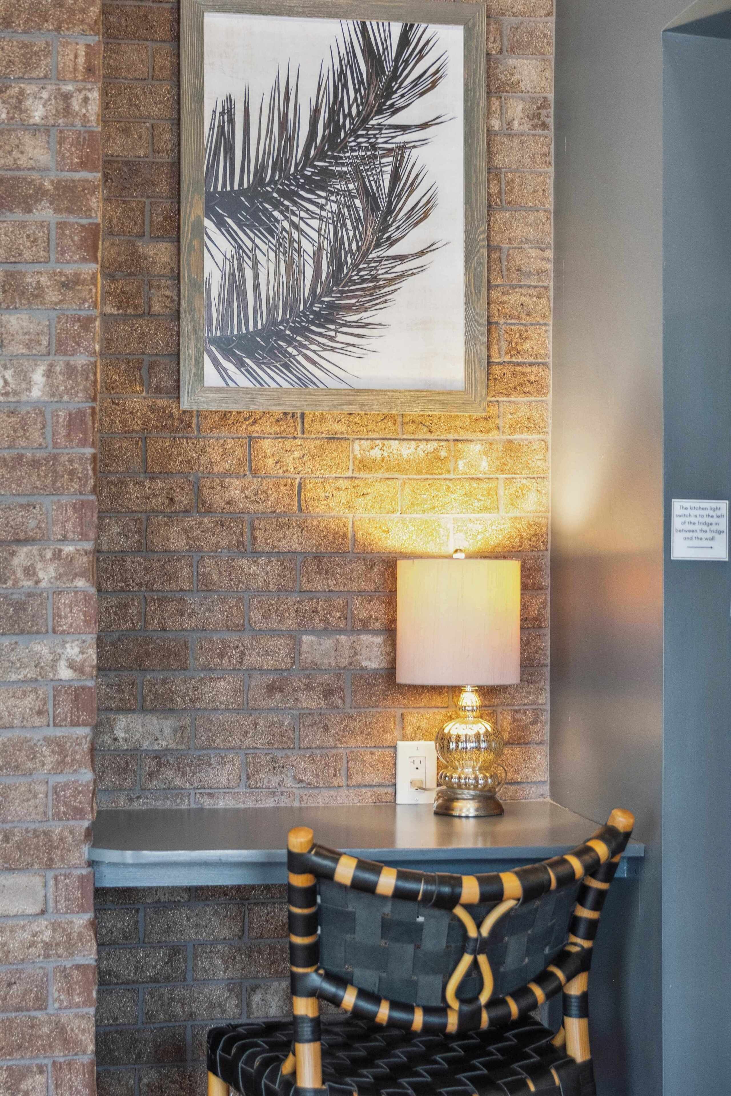 A brick wall with a framed leaf artwork, a table with a lamp, and a wicker chair.