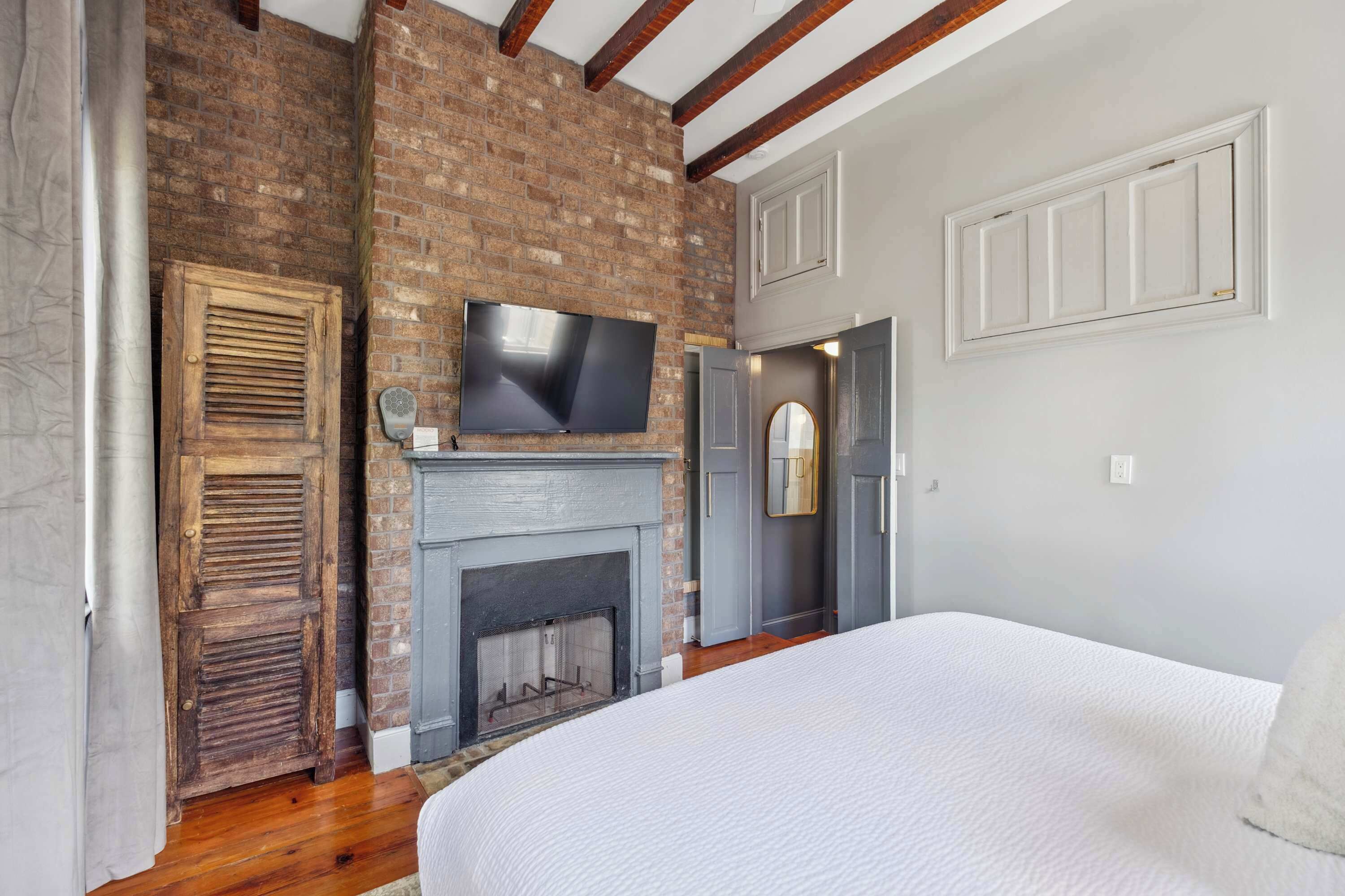 Un dormitorio con pared de ladrillo, chimenea, televisor montado encima y cama con cubrecama blanco.