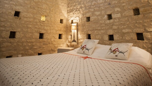 Una habitación con techo alto, vigas de madera a la vista, paredes de piedra, una cama con colcha estampada y un tragaluz.