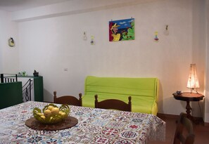 Une pièce avec un lit, une table sur laquelle se trouve un panier de fruits, une lampe et un tableau accroché au mur.