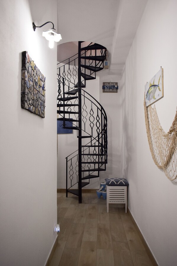 Un escalier en colimaçon avec une rampe en métal noir, une œuvre d’art fixée au mur, une enseigne « Relax Beach » et une décoration murale en macramé.