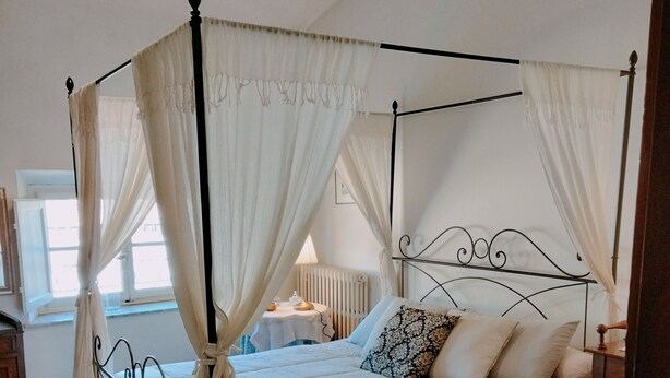 A four-poster canopy bed with a white bedspread and matching curtains, a wooden nightstand with a lamp, and a view of the room's wooden floor and a window with a white curtain. - Talents Home Historical center (Lucca)