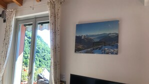 Un salon avec un téléviseur, une fenêtre avec des rideaux et un tableau représentant un paysage de montagne.