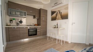 Eine moderne Küche mit Holzschränken, einem eingebauten Backofen und einer weißen Arbeitsplatte. Ein kleiner weißer Hocker und ein an der Wand befestigtes Regal mit Flasche und Glas sind ebenfalls vorhanden.