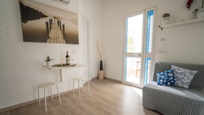 Ein modernes Wohnzimmer mit einem grauen Sofa, zwei weißen Hocker, einem kleinen Tisch mit Flasche und Glas sowie einem großen Gemälde eines Piers.