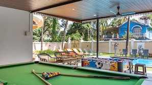 A pool table with a foosball table, pool cues, and balls, set in a covered area with a view of a pool and lounge chairs. - Villa Calangute Phase 4, Near Calangute Beach & Restaurants (Calangute)