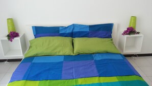 Una cama con una colcha azul y verde a cuadros, flanqueada por mesitas de noche blancas con jarrones verdes y flores púrpuras.