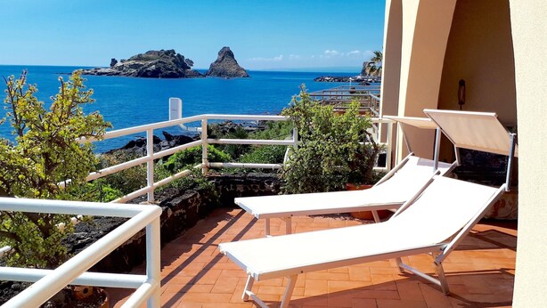 Una terraza con sillones blancos, baranda y vista al mar y formaciones rocosas.
