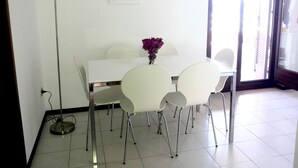 Un comedor con una mesa blanca, cuatro sillas blancas, un jarrón con flores moradas, una lámpara de pie y un cuadro en la pared.
