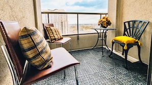 A balcony with two chairs, one with a plaid cushion and another with a yellow cushion, a small table with a vase of flowers, and a view of buildings through a large window. - 2-Bed, 2-Bath Condo w/Pool & Parking by Square 1 (Mississauga)