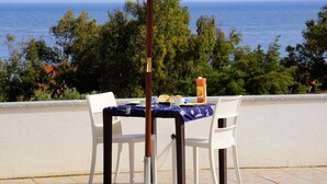 Een tafel met een blauwe tafelkleed en witte stoelen, een fles sap en een beker, voor een achtergrond van een helderblauwe lucht en de zee.