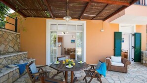 A covered patio with a stone floor, a table set for dining, and a seating area with chairs and a sofa. - Olga Luxury Apt Lower ( Adults only) (Paxoi)