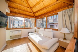 A cozy room with a wooden ceiling, a sofa, a TV, and a window with a view of buildings.