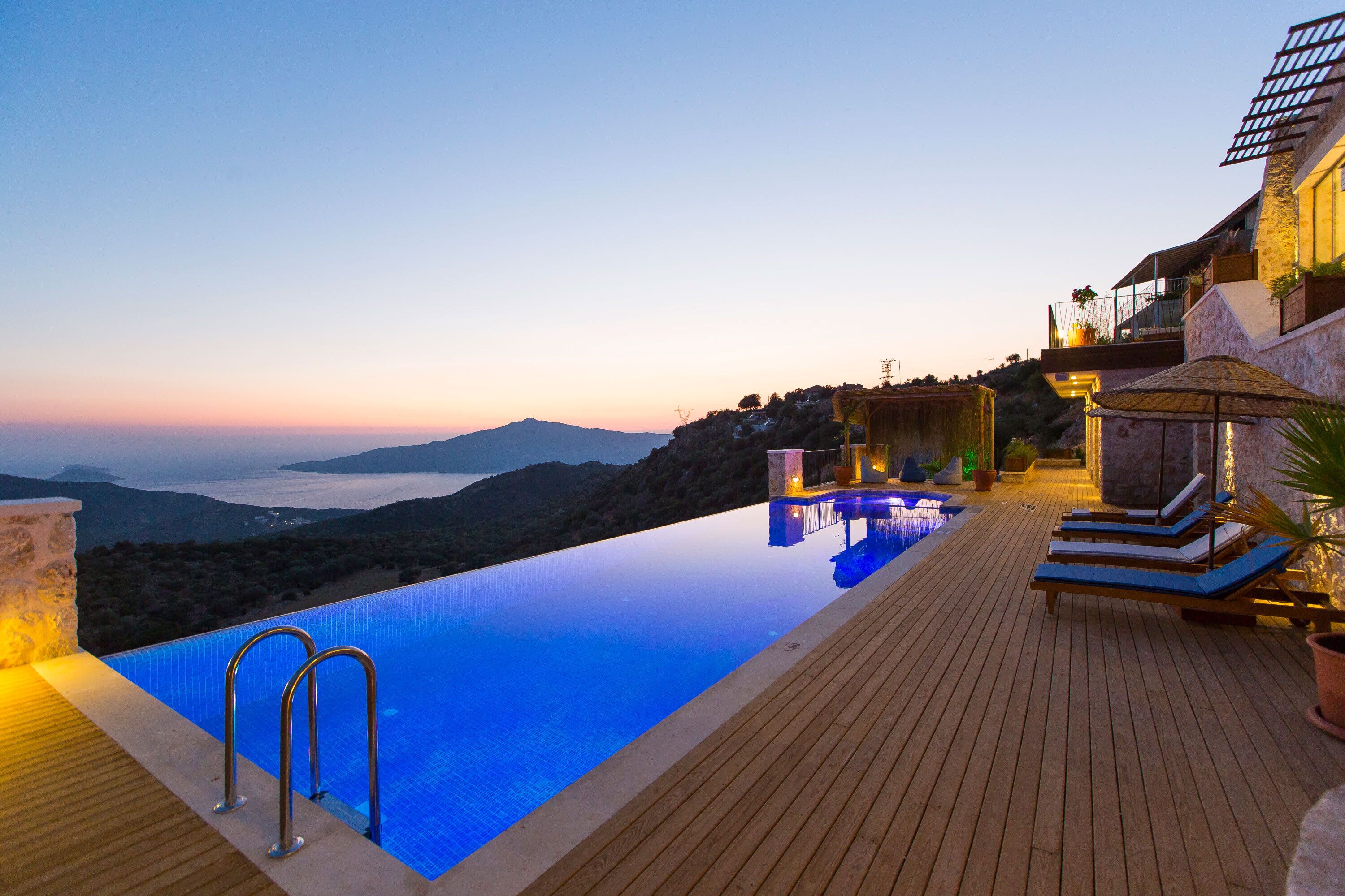 A infinity pool with wooden deck and lounge chairs, overlooking a mountainous landscape.