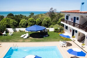 A pool area with a view of the sea, outdoor seating, and a building with a balcony. - Pousada das Flores (Anchieta)