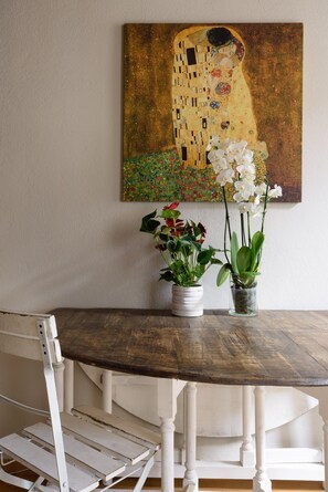 Una mesa de comedor de madera con sillas blancas, un cuadro de una mujer y un niño, y dos macetas con orquídeas.