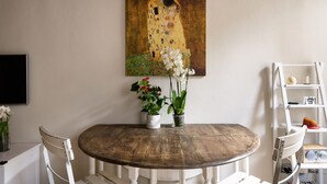 Un comedor con una mesa redonda de madera, sillas blancas, un cuadro en la pared y dos macetas con flores.