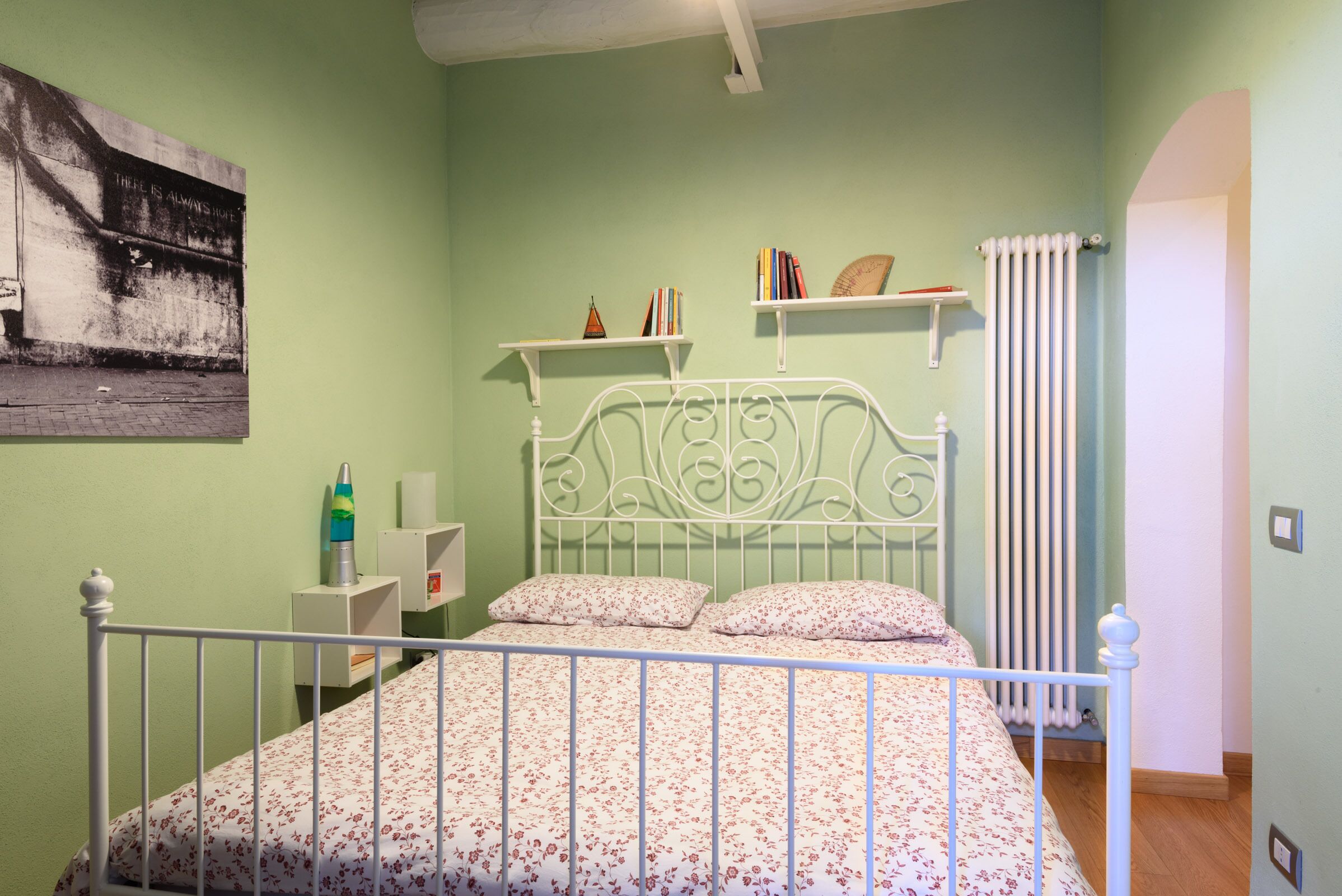 A bedroom with a bed, a radiator, and shelves with books and decorative items.