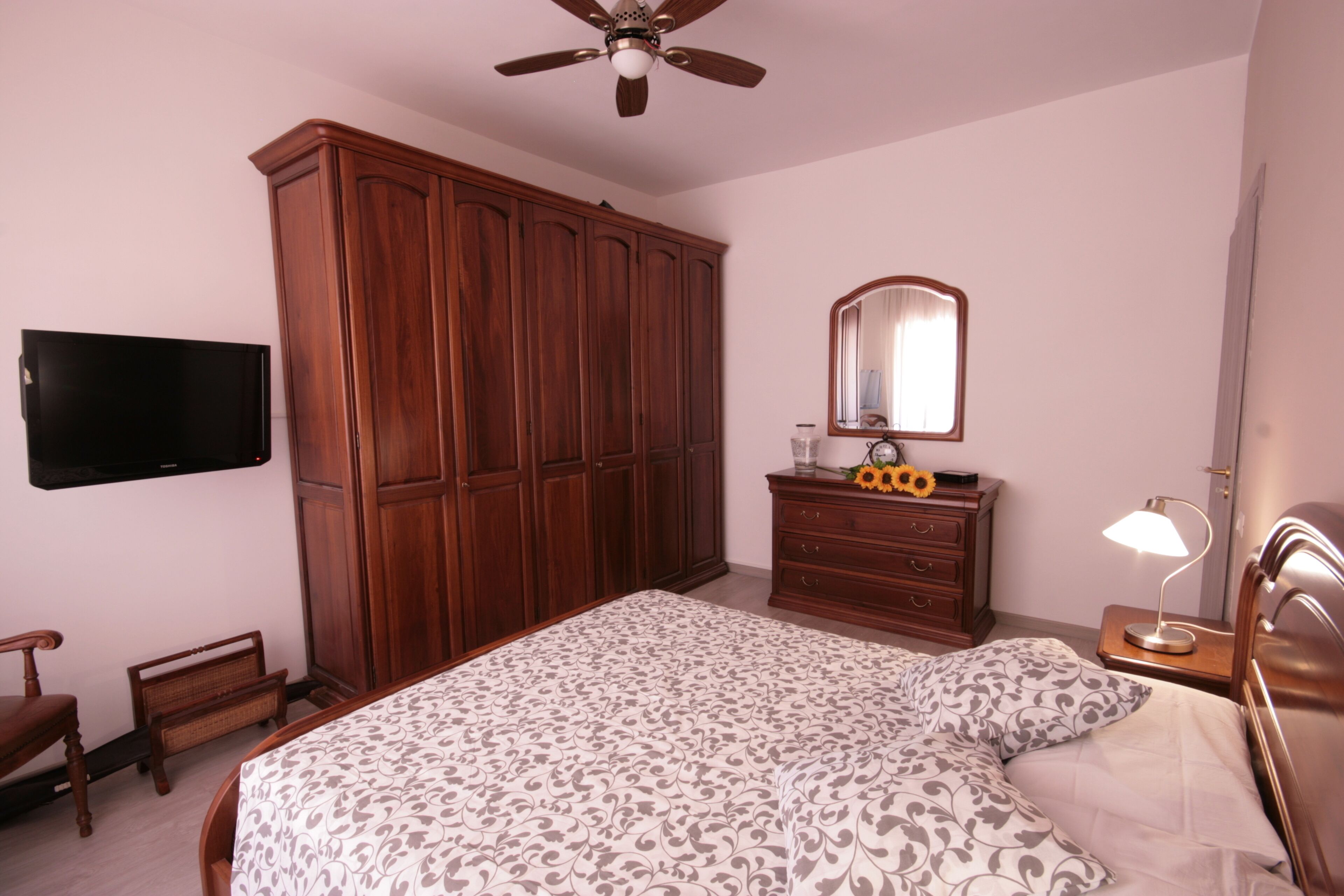 A bedroom with a large bed, wooden wardrobe, a television, a dresser, a mirror, and a ceiling fan.