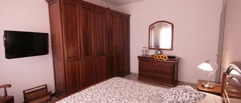 A bedroom with a large bed, wooden wardrobe, a television, a dresser, a mirror, and a ceiling fan.