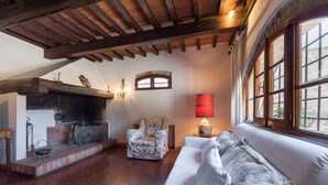 A cozy living room with a fireplace, a floral armchair, a white sofa, and large windows.