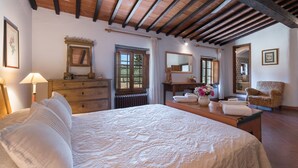 A bedroom with a large bed, wooden ceiling beams, a dresser, a mirror, and a chair.