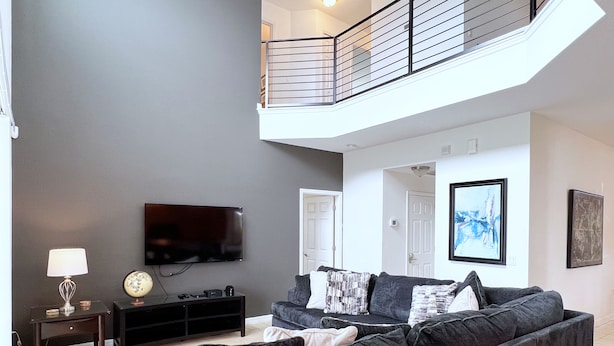 A modern living room with a grey sofa, a flat-screen TV, a small table with a lamp, and a staircase leading to an upper level.
